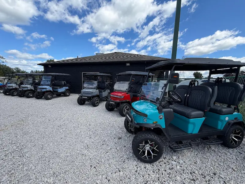 A line up multi seated golf carts on a gravel parking lot with clouds in the sky.